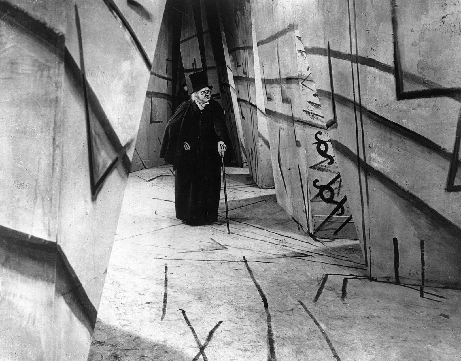 A frame from 'The Cabinet of Dr. Caligari' showing the style of German Expressionism, with a stark contrast and an erratic and angular mise-en-scene, Dr. Caligari walks through a long corridor, wearing a hat, suit and cane